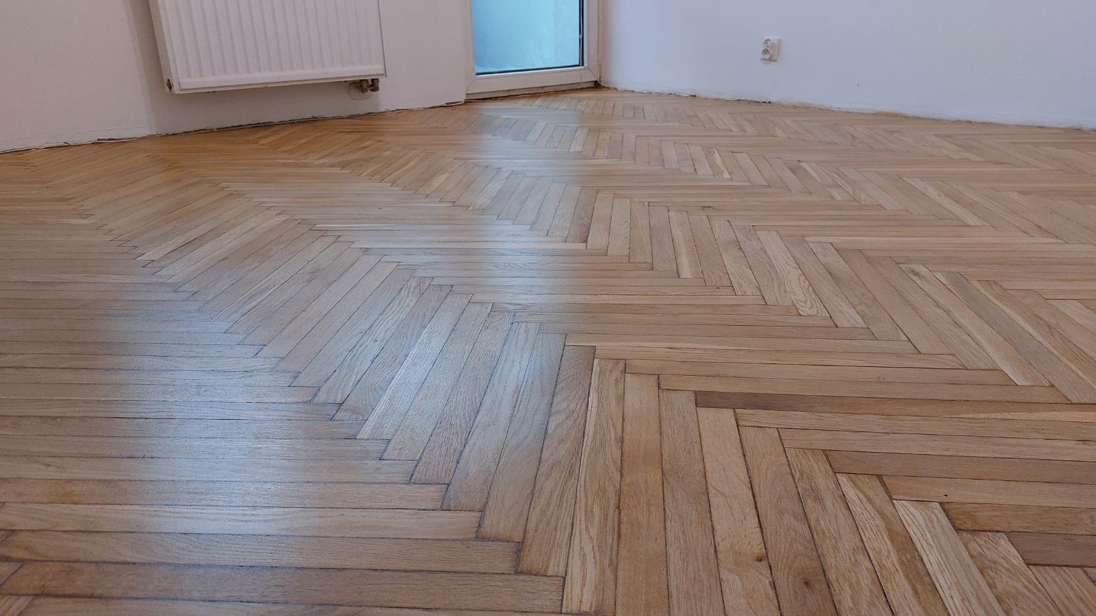 Ułożony parkiet w jodełkę, jasne drewno, widok z góry, fragment ściany z grzejnikiem i okno w tle. Układanie parkietu z dbałością o detale.