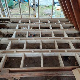 Gravida Build - Drewniana konstrukcja podłogi w trakcie budowy, widoczne legary i przestrzeń między nimi. W tle okno z widokiem na zewnątrz i narzędzia budowlane.