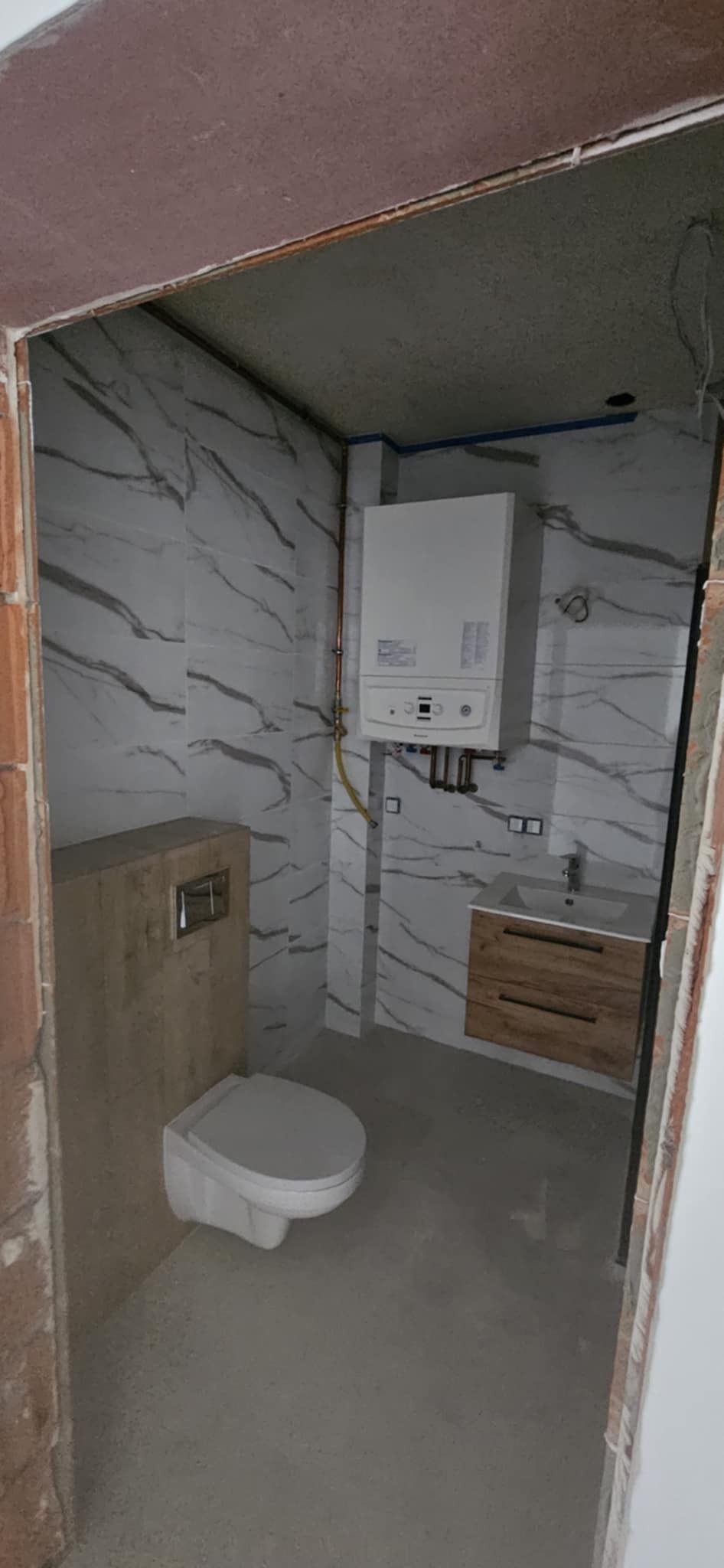 Wykończona łazienka z marmurowymi płytkami, wiszącą toaletą, szafką z umywalką i piecem gazowym. Widok zza futryny, surowy stan wykończenia.