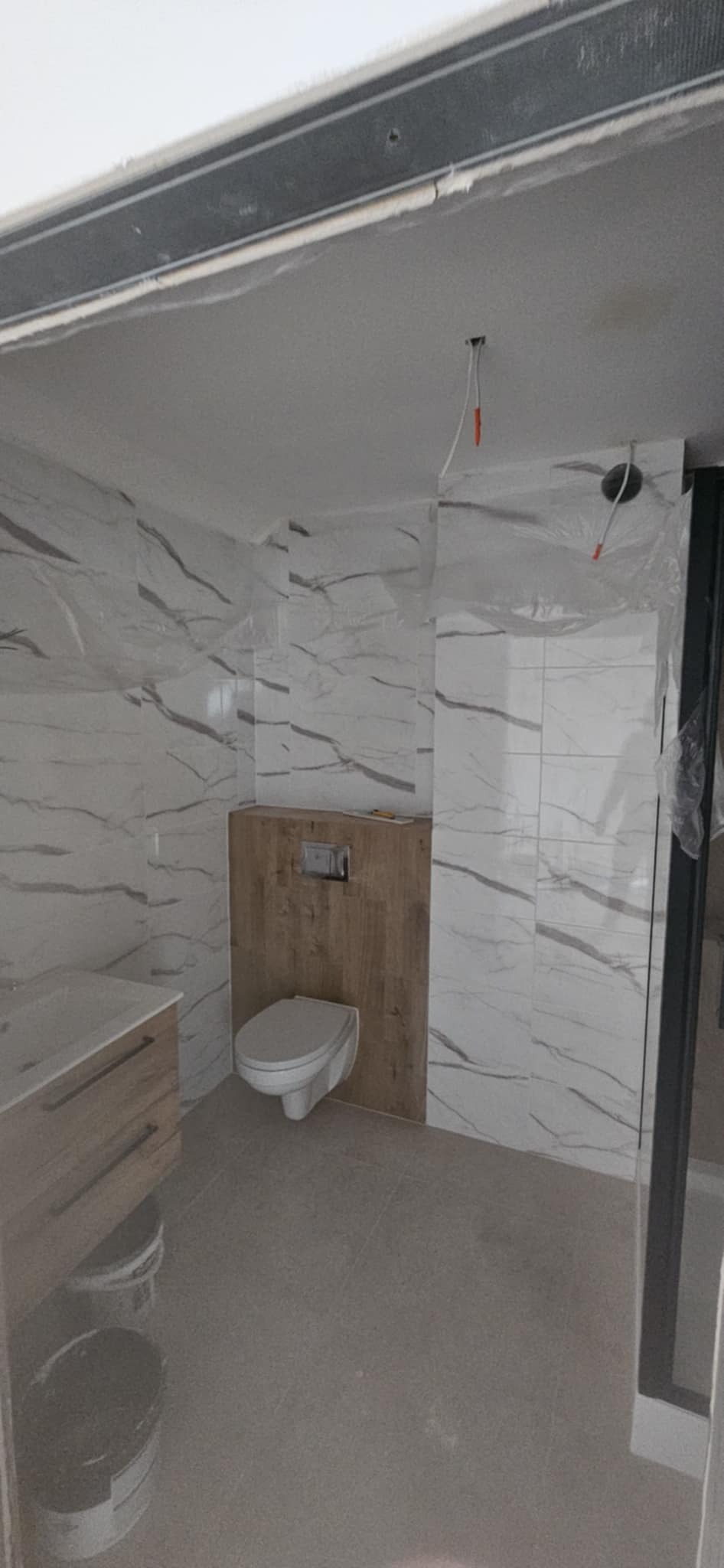 Wnętrze łazienki w trakcie wykańczania: białe płytki imitujące marmur, drewnopodobna zabudowa WC, częściowo widoczna szafka z umywalką, szare płytki podłogowe.