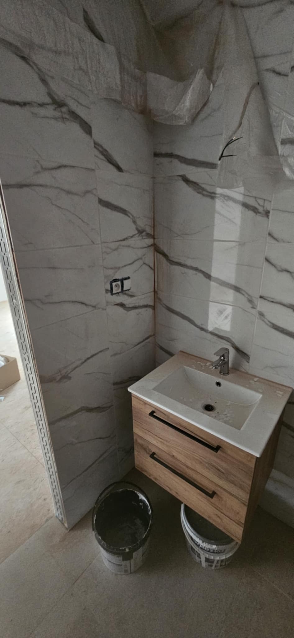 Wykończona łazienka z marmurowymi płytkami i drewnianą szafką pod umywalką. Widoczne wiaderka po farbie, sugerujące świeży remont. Minimalistyczny design.