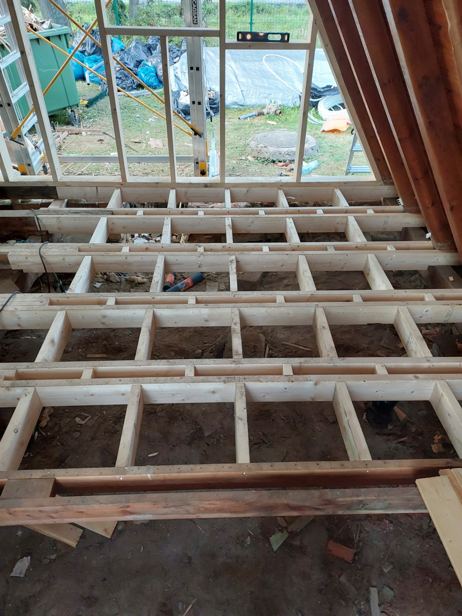 Drewniana konstrukcja podłogi w trakcie budowy, widoczne legary i przestrzeń między nimi. W tle okno z widokiem na zewnątrz i narzędzia budowlane.