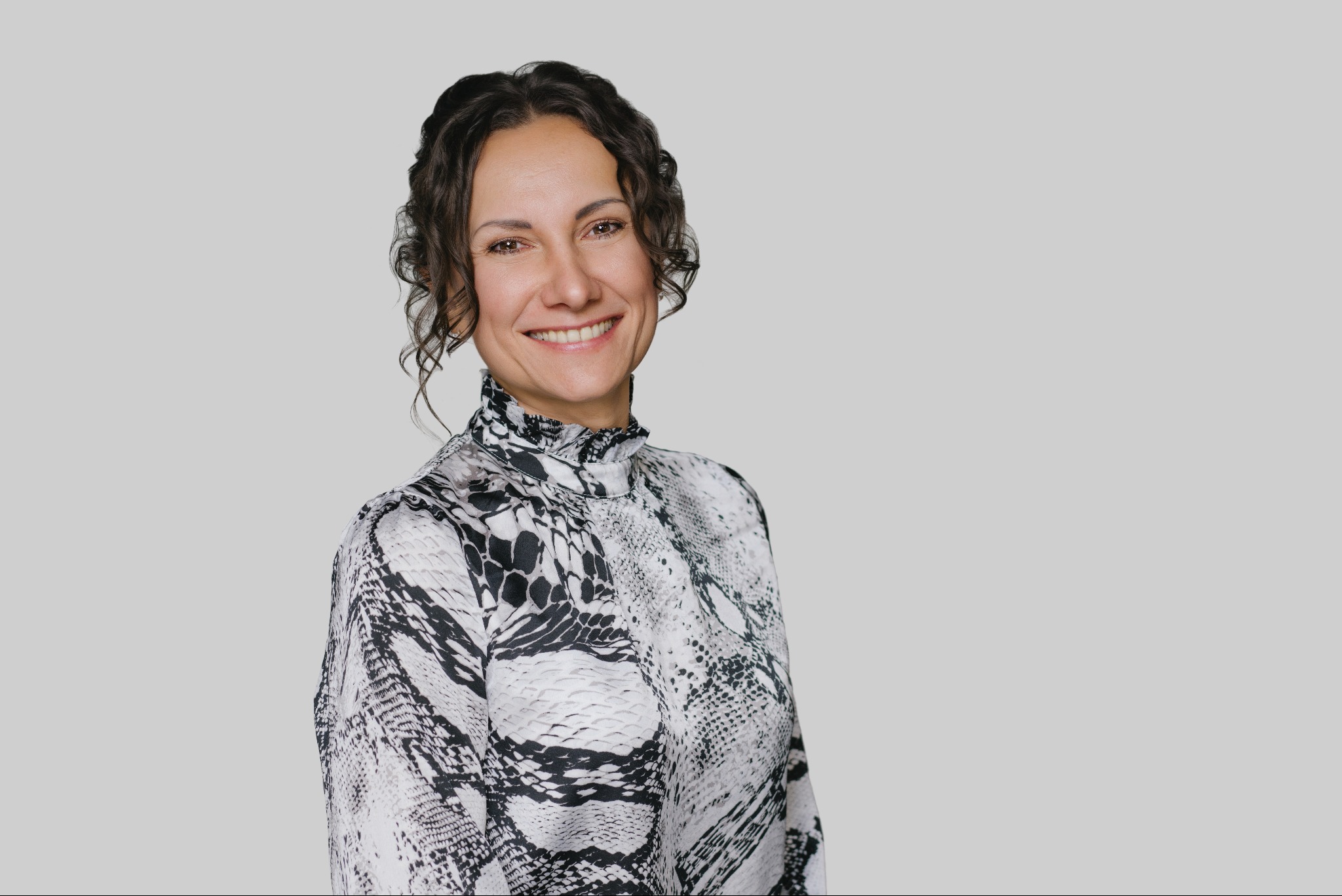 Uśmiechnięta brunetka w bluzce we wzór skóry węża, na jasnoszarym tle. Portret agenta nieruchomości, budzący zaufanie i profesjonalizm.