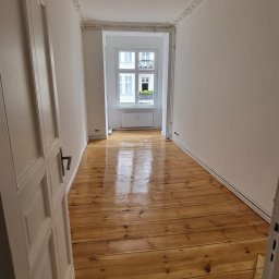 Jasne, odnowione deski podłogowe z widocznym połyskiem w pustym pokoju z białymi ścianami i oknem. Częściowo otwarte białe drzwi po lewej stronie kadru.