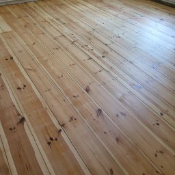 Jasna, drewniana podłoga z widocznymi słojami, po cyklinowaniu, w pustym pokoju. Widoczne listwy przypodłogowe i fragment ściany. Ujęcie z niskiego kąta.