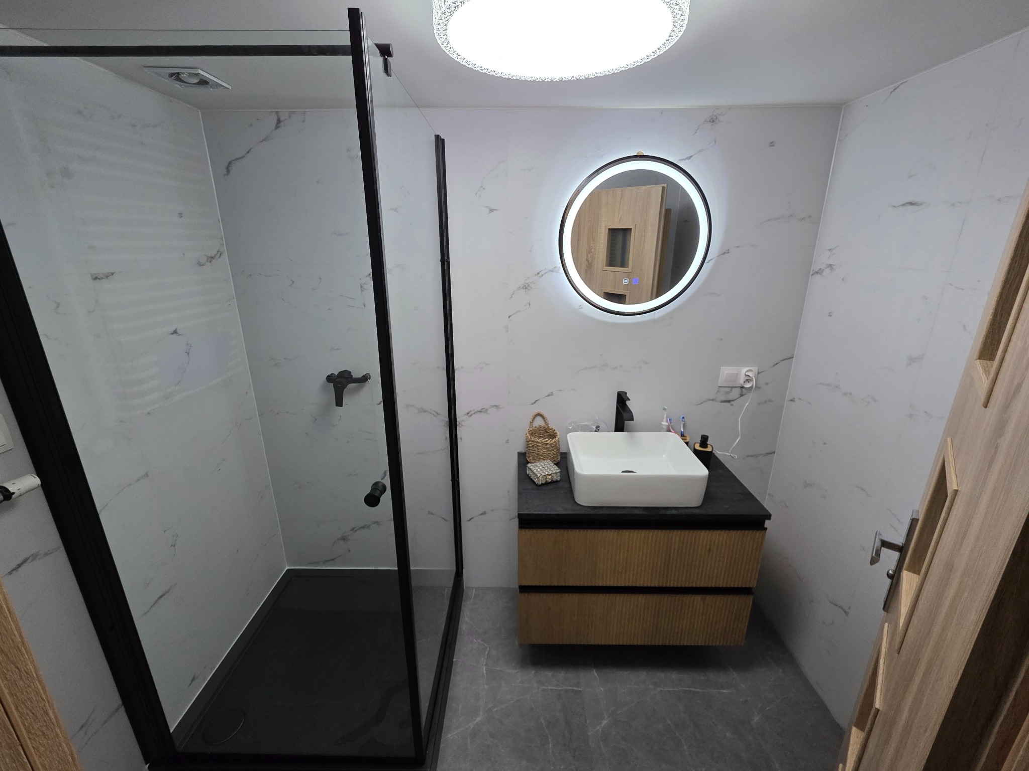 Nowoczesna łazienka z kabiną prysznicową typu walk-in, umywalką nablatową na szafce z lamelami i okrągłym lustrem z podświetleniem LED. Białe płytki imitujące marmur.