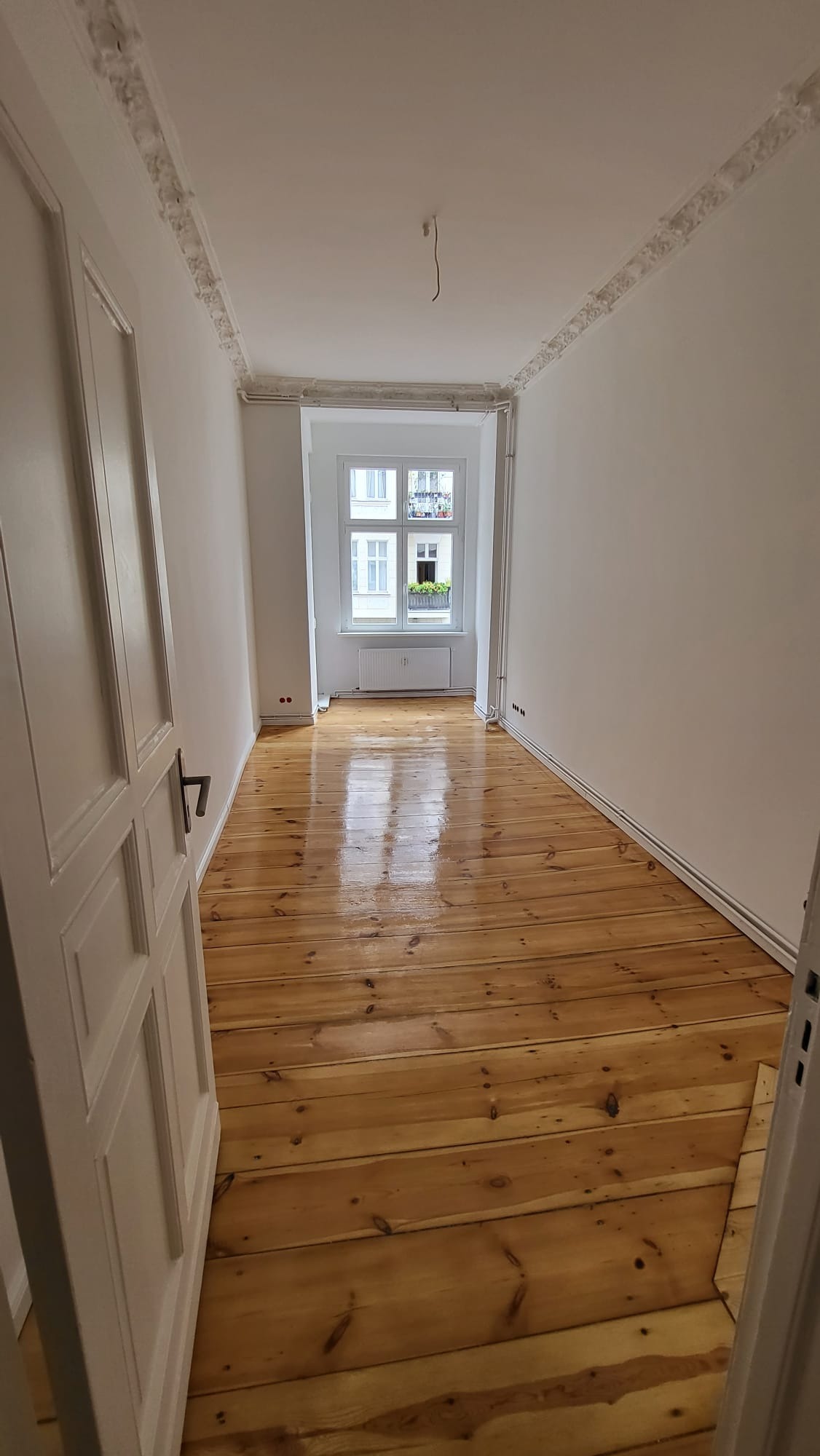 Jasne, odnowione deski podłogowe z widocznym połyskiem w pustym pokoju z białymi ścianami i oknem. Częściowo otwarte białe drzwi po lewej stronie kadru.