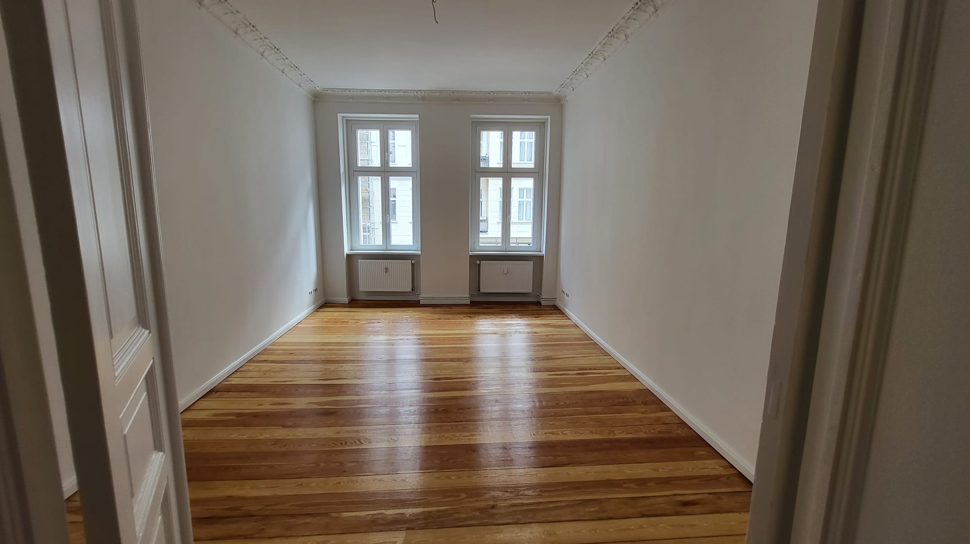 Pusty pokój po remoncie z drewnianą podłogą, białymi ścianami i dwoma oknami. Widok zza otwartych drzwi, listwy dekoracyjne pod sufitem.