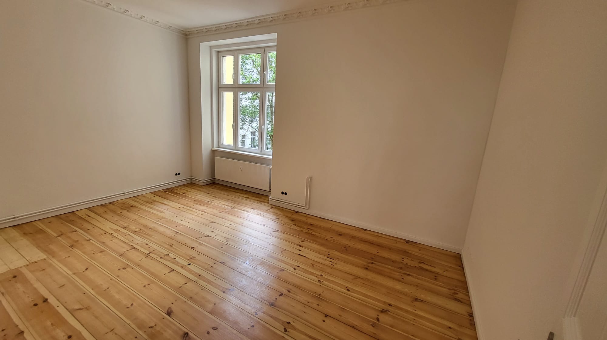 Jasne, puste wnętrze z drewnianą podłogą i oknem. Świeżo pomalowane ściany i listwy przypodłogowe. Widoczny grzejnik pod oknem i instalacja grzewcza przy ścianie.