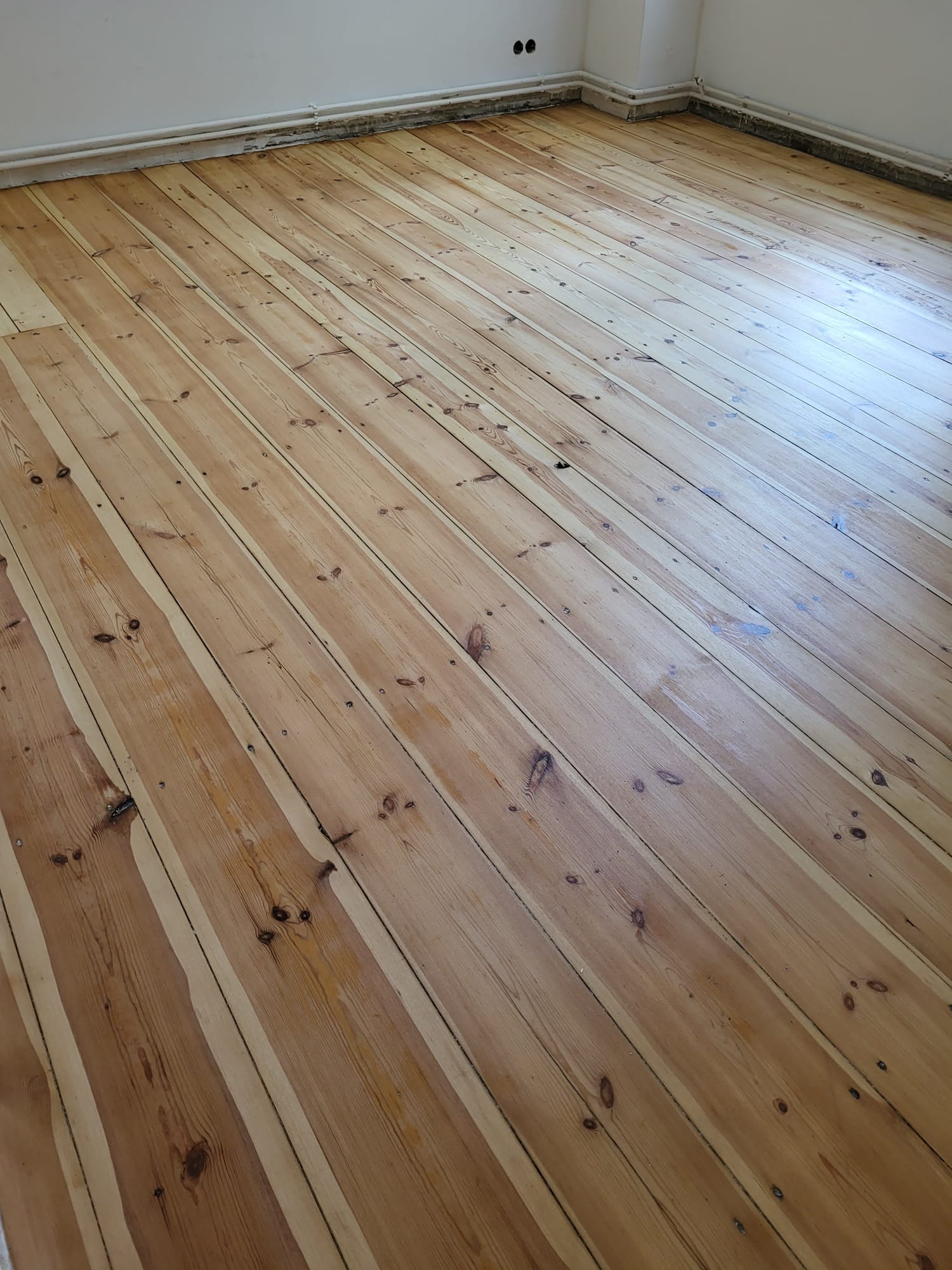 Jasna, drewniana podłoga z widocznymi słojami, po cyklinowaniu, w pustym pokoju. Widoczne listwy przypodłogowe i fragment ściany. Ujęcie z niskiego kąta.