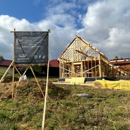 PYTON WOOD - Wznoszenie szkieletu domu z drewna w słoneczny dzień. Na pierwszym planie tablica reklamowa firmy budowlanej z listą usług. Widoczne materiały budowlane.