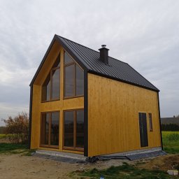 PYTON WOOD - Żółty dom z drewnianą elewacją i czarnym dachem, duże okna, komin. Widok z zewnątrz, teren budowy, butelka wody i elementy konstrukcyjne w tle.