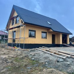 PYTON WOOD - Surowy stan domu w budowie z czarnym dachem i żółtymi ścianami, otoczony rusztowaniami i stertą drewna na błotnistym terenie w pochmurny dzień.