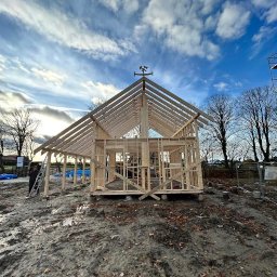 PYTON WOOD - Wznoszenie konstrukcji szkieletowej domu z drewnianą pogodynką na szczycie dachu, na tle błękitnego nieba z chmurami. Widoczny teren budowy i elementy rusztowania.
