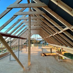 PYTON WOOD - Wnętrze domu w trakcie budowy, widoczna drewniana konstrukcja dachu i ścian, z zewnątrz widać zieleń i zabudowania. Jasne, naturalne światło.