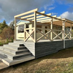 PYTON WOOD - Jasny taras z kompozytu z drewnianą konstrukcją pergoli przy domu. Schody prowadzą na podest z balustradą. Błękitne niebo z chmurami w tle.