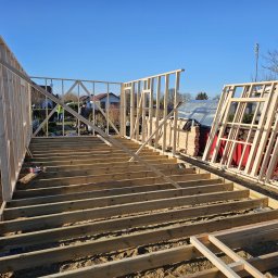 PYTON WOOD - Wczesny etap budowy domu: drewniany szkielet z widoczną podłogą i fragmentami ścian, na tle ogrodu i błękitnego nieba. Widoczne detale konstrukcyjne.