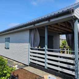 PYTON WOOD - Szary dom modułowy z zadaszonym tarasem i zasłonami, widok z zewnątrz w słoneczny dzień. Widoczne detale wykończenia i otoczenie domu.