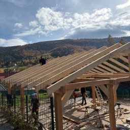 PYTON WOOD - Montaż drewnianej konstrukcji dachu na tle górskiego krajobrazu. Widoczne elementy więźby i pracownicy podczas pracy na wysokości.