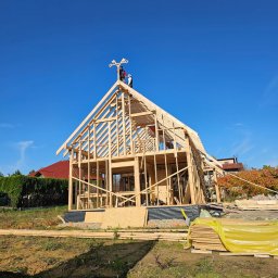 PYTON WOOD - Wznoszenie konstrukcji domu z drewna, widoczna więźba dachowa i pracownicy na szczycie. Błękitne niebo w tle. Elementy konstrukcyjne na ziemi.