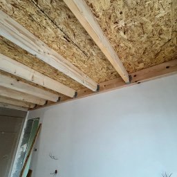 PYTON WOOD - Surowe drewniane belki stropowe z płytą OSB, widok z dołu w trakcie budowy. Metalowe łączniki, białe ściany w tle, widoczne przewody elektryczne.