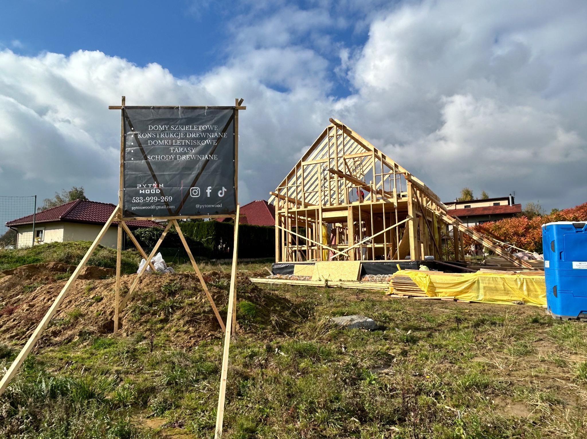 Wznoszenie szkieletu domu z drewna w słoneczny dzień. Na pierwszym planie tablica reklamowa firmy budowlanej z listą usług. Widoczne materiały budowlane.