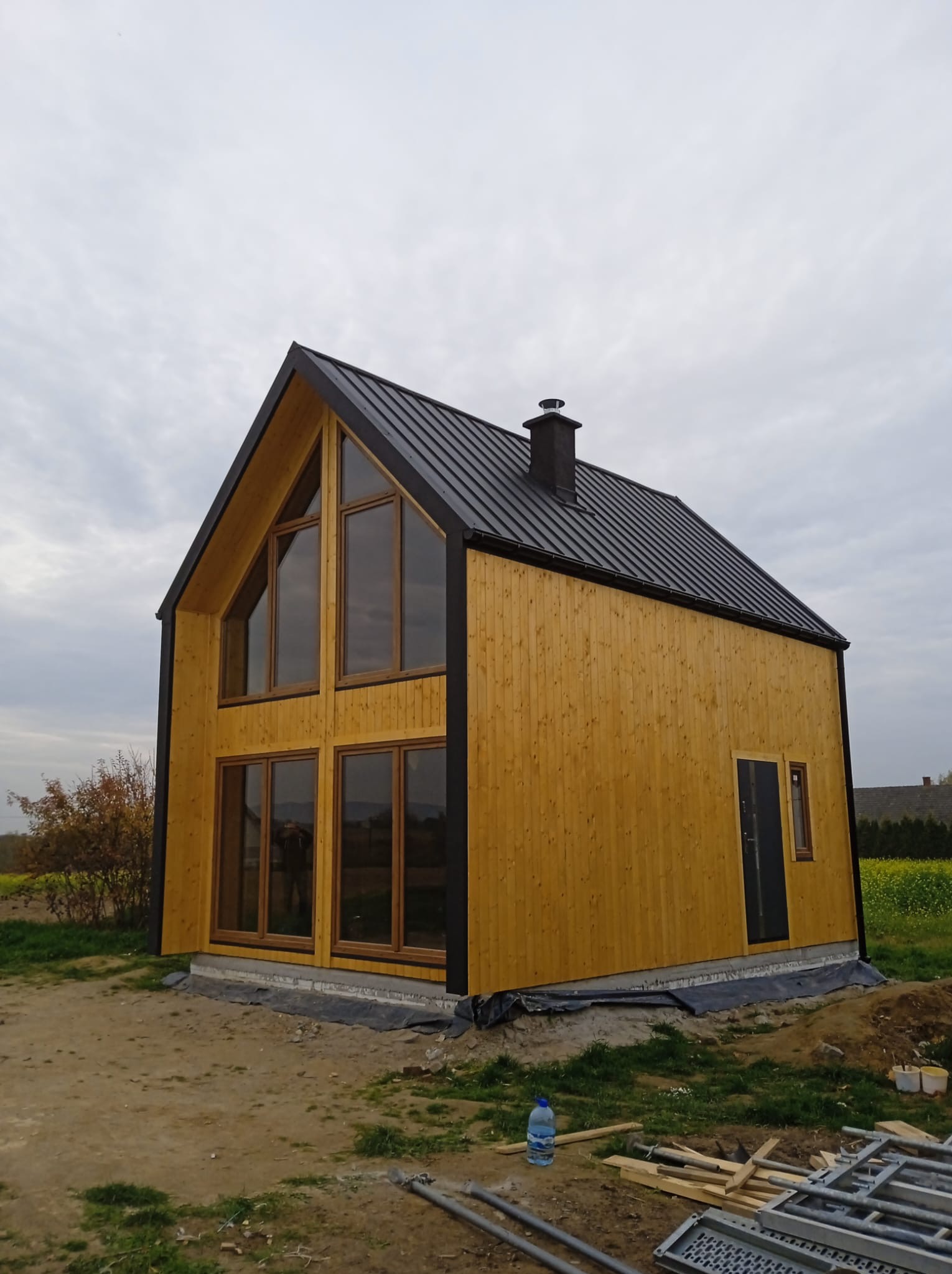 Żółty dom z drewnianą elewacją i czarnym dachem, duże okna, komin. Widok z zewnątrz, teren budowy, butelka wody i elementy konstrukcyjne w tle.
