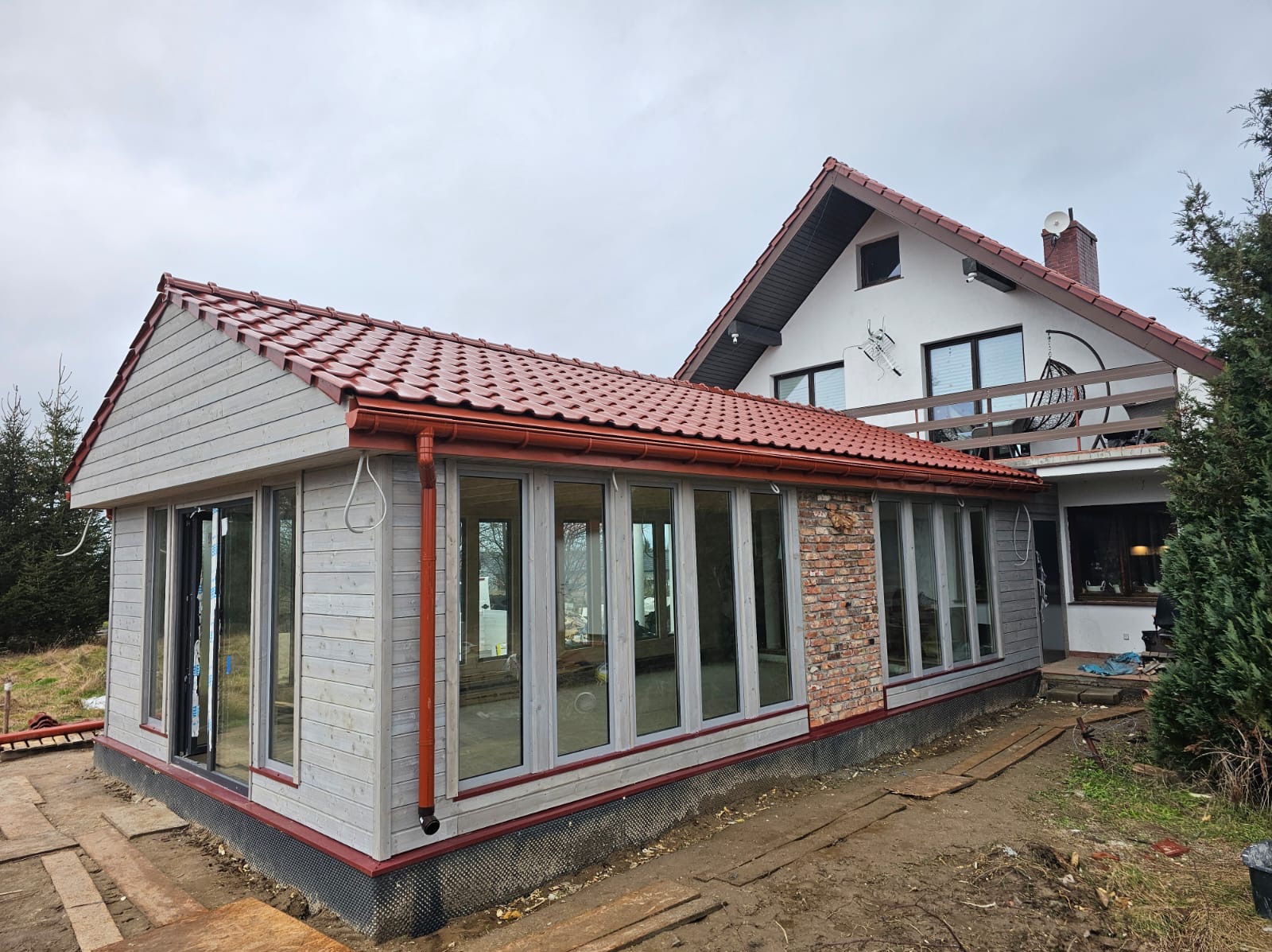 Dobudowana przeszklona weranda z drewnianą elewacją i czerwoną dachówką, przylegająca do domu z balkonem. Widoczne elementy wykończeniowe i teren wokół w trakcie prac.