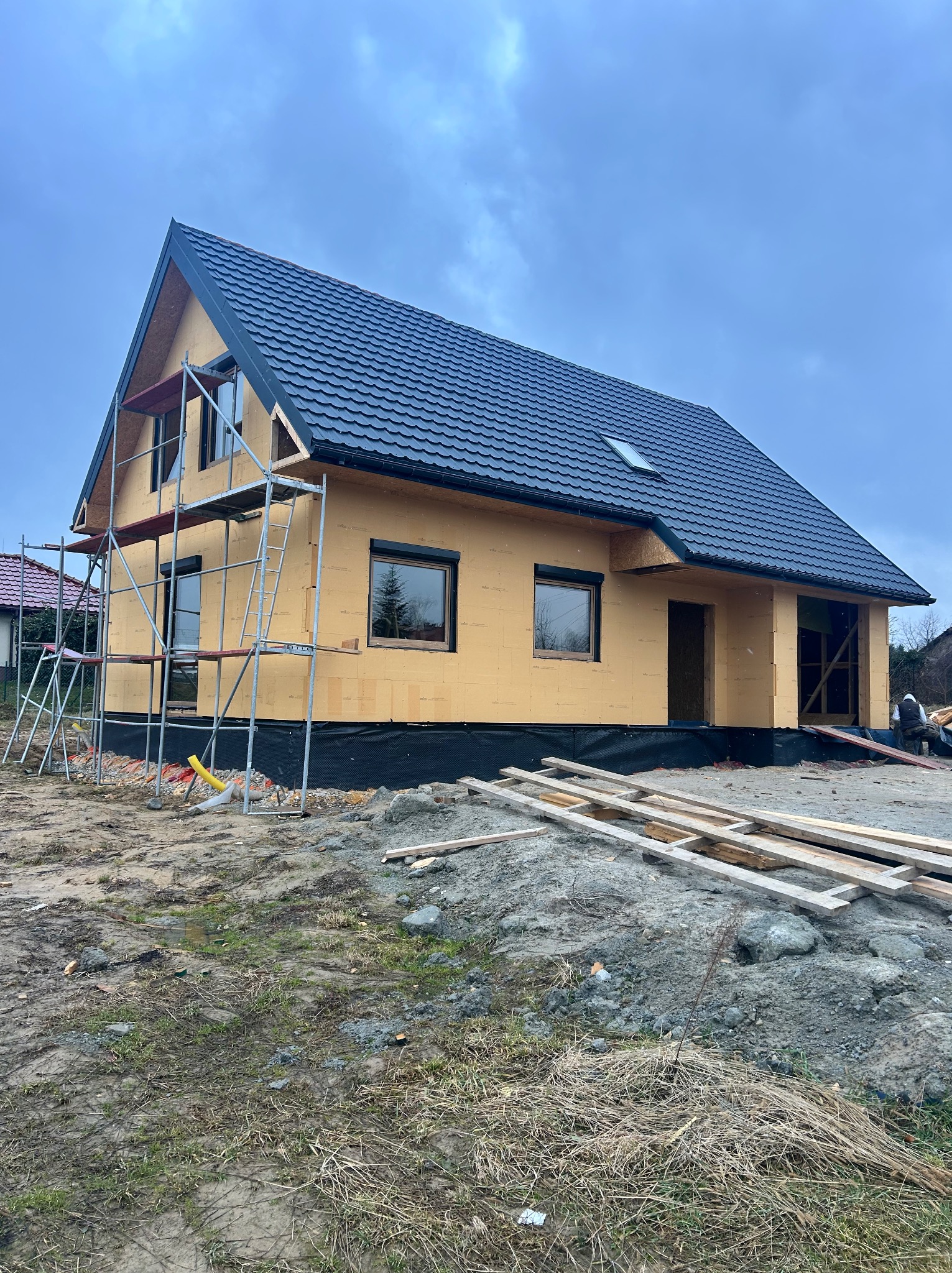 Surowy stan domu w budowie z czarnym dachem i żółtymi ścianami, otoczony rusztowaniami i stertą drewna na błotnistym terenie w pochmurny dzień.