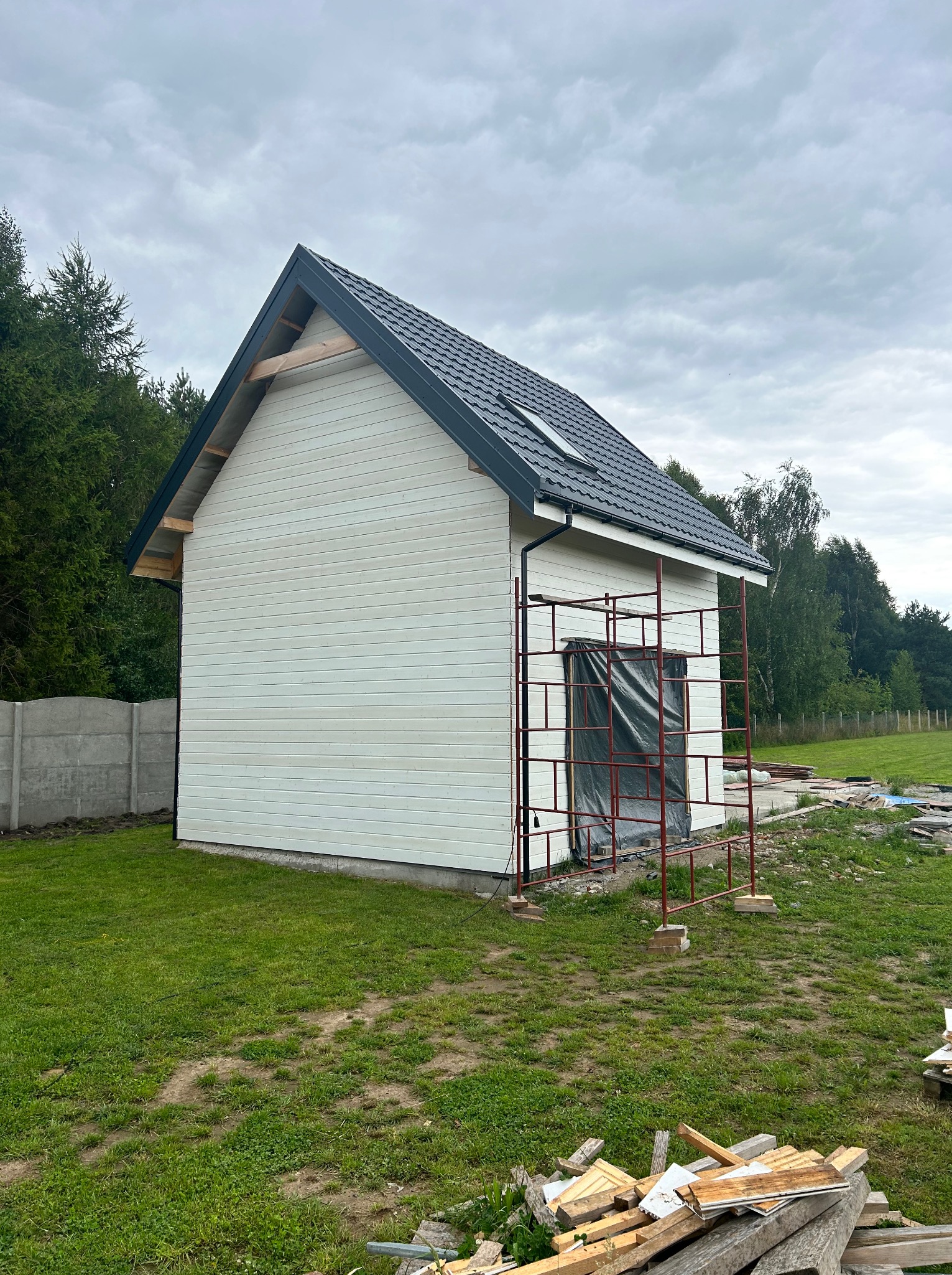 Boczny widok na biały dom z ciemnym dachem w trakcie budowy, z widocznymi rusztowaniami i elementami wykończeniowymi. W tle zieleń drzew i betonowe ogrodzenie.