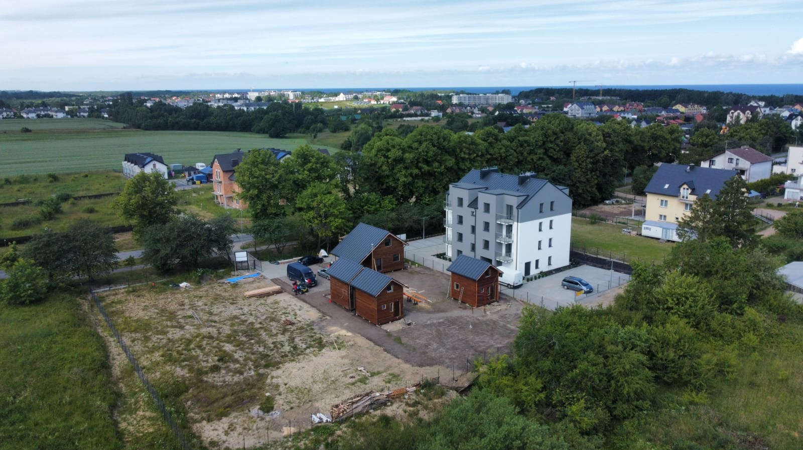 Widok z lotu ptaka na plac budowy z trzema małymi, brązowymi domkami z ciemnymi dachami i białym budynkiem apartamentowym w tle, otoczonym zielenią i zabudową miejską.