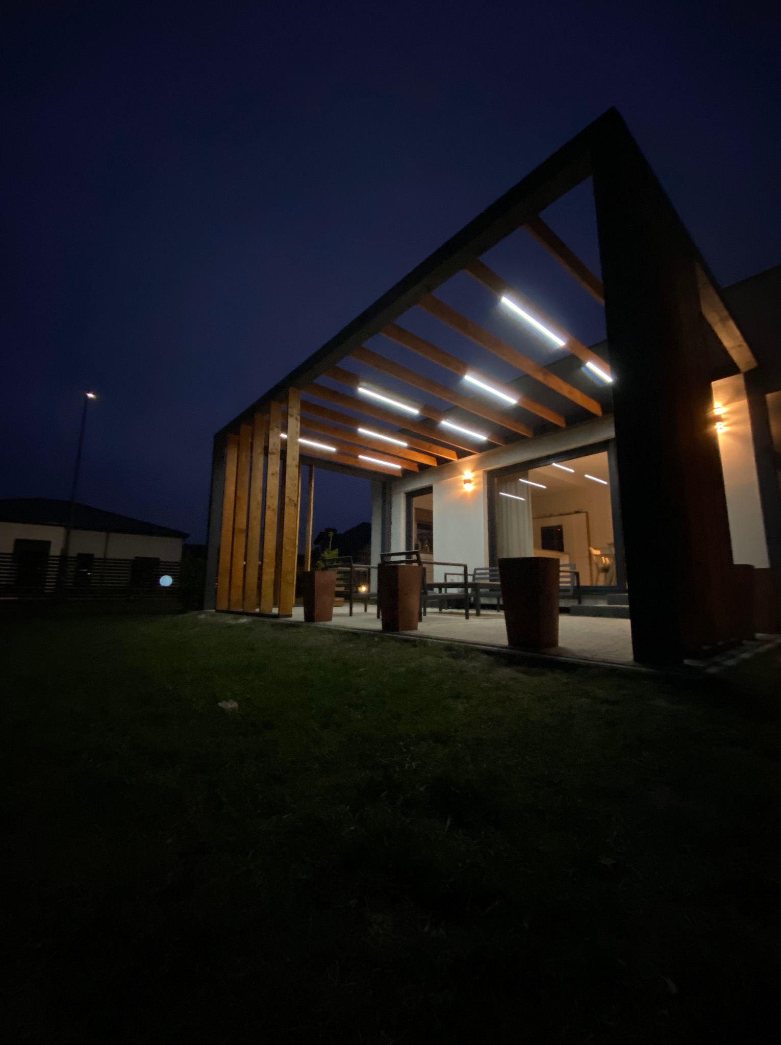 Nowoczesna pergola z drewnianą konstrukcją i oświetleniem LED, widoczna nocą. Minimalistyczny design, ciemne tło nieba, oświetlony taras z meblami.