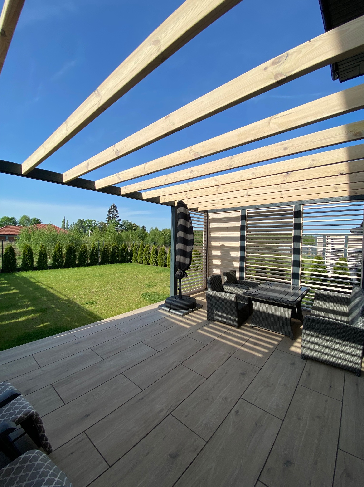 Nowoczesna pergola z jasnego drewna nad tarasem z szarymi płytkami. W tle zielony trawnik i rząd tuj. Meble ogrodowe z rattanu.