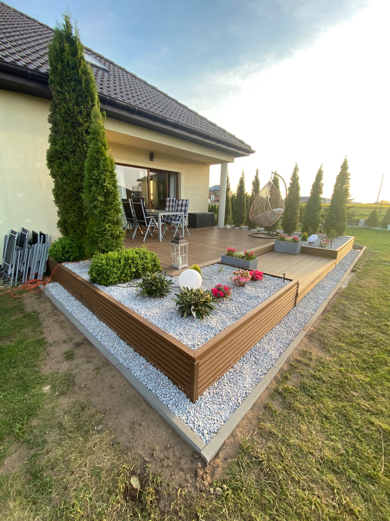 Nowoczesny taras z desek kompozytowych, otoczony rabatami z białego żwiru i roślinnością. W tle dom z meblami ogrodowymi i wiszącym fotelem.