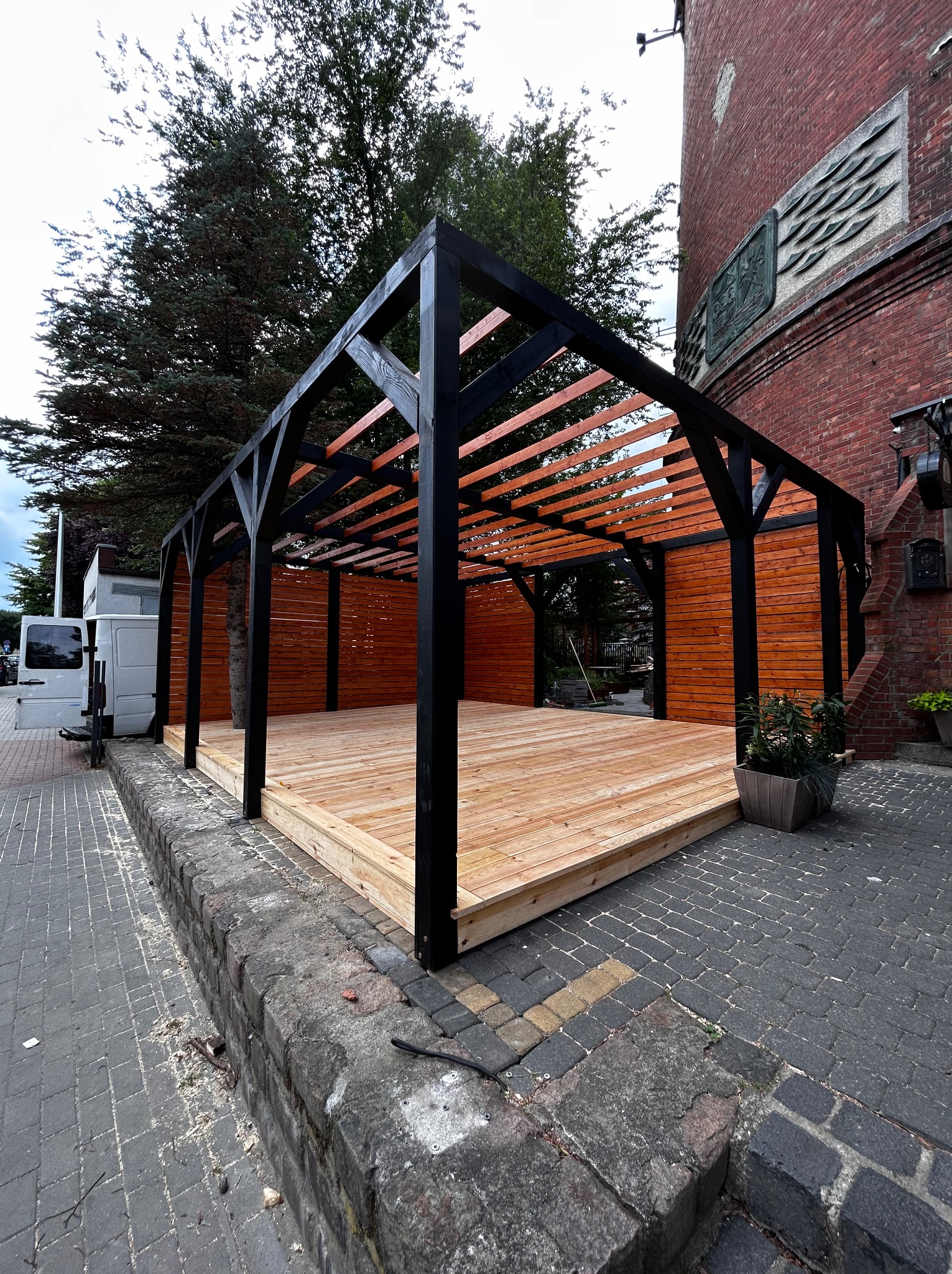 Nowoczesna pergola z drewna, czarne słupy i pomarańczowe deski, na tle ceglanego budynku i zieleni. Widoczne ślady obróbki drewna.