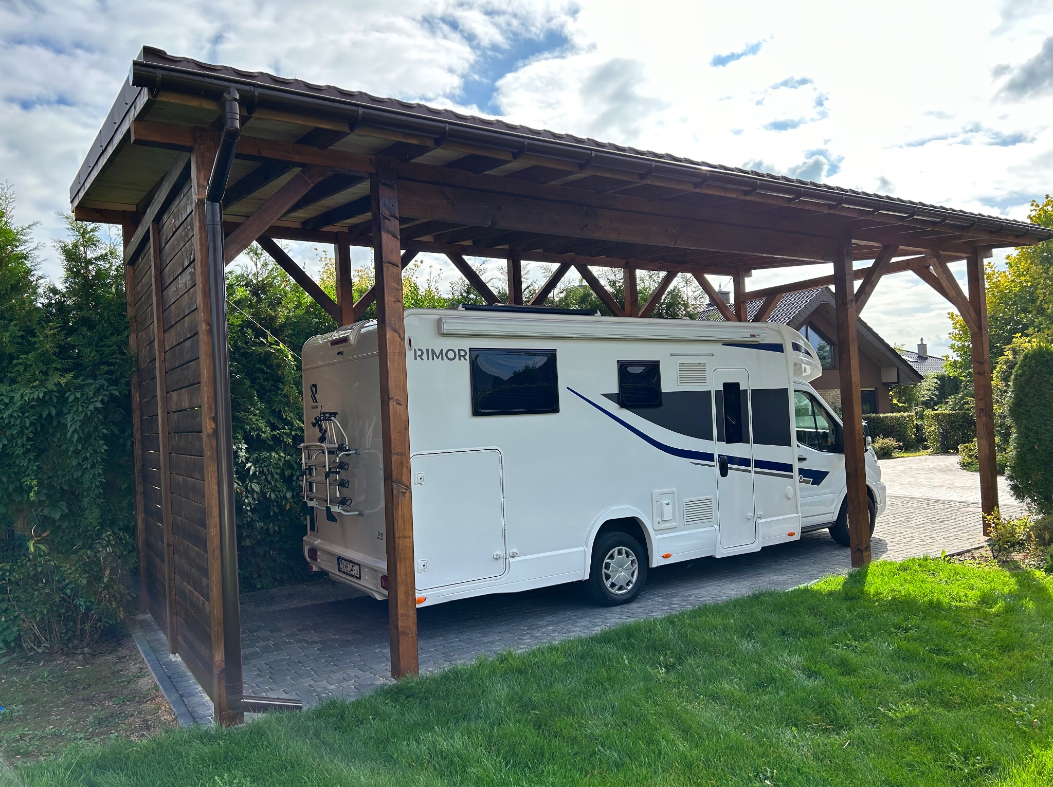 Drewniana wiata garażowa z zadaszeniem, chroniąca biały kamper. Konstrukcja z belek, częściowo zabudowana deskami, otoczona zielenią. Widok z boku, w słoneczny dzień.