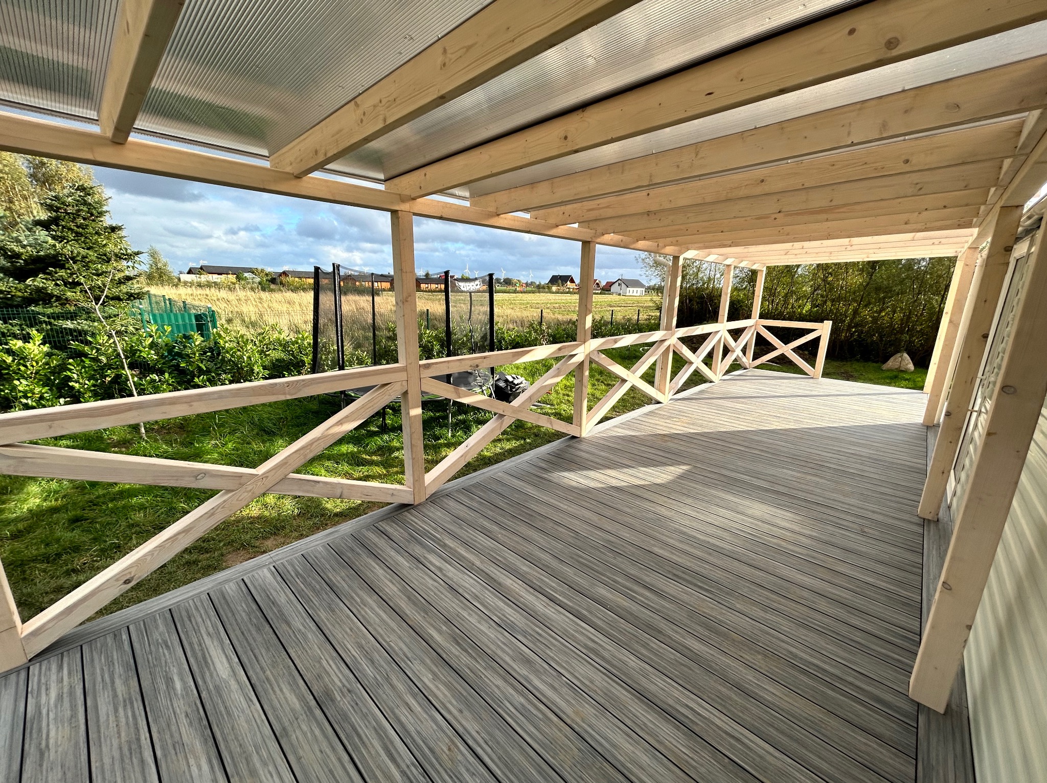 Zadaszony taras z szarej deski kompozytowej i drewnianą balustradą krzyżową. Widok na zielony ogród i zabudowania w oddali. Lekkie zachmurzenie.