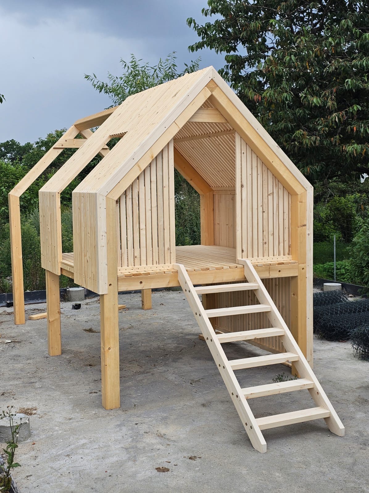 Surowa, drewniana konstrukcja domku na palach z drabinką, w trakcie budowy. Widoczne elementy więźby dachowej i deski elewacyjne. Tło z zielenią.