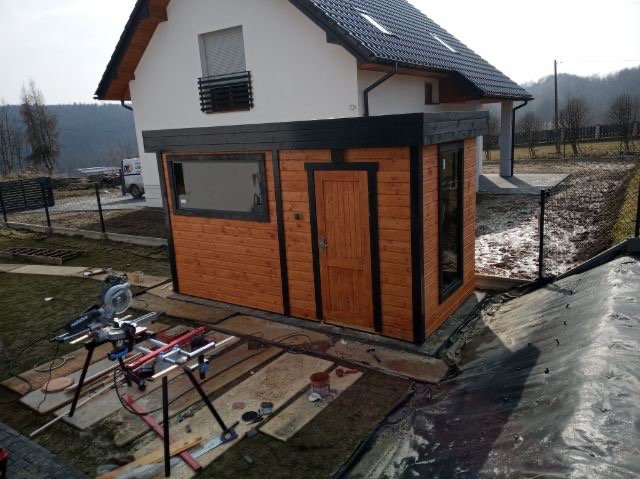 Drewniana dobudówka z oknami i drzwiami, pomalowana na brązowo z czarnymi obramowaniami, przylegająca do białego domu. W tle widoczne drzewa i narzędzia pracy.