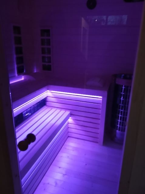 Nowoczesna sauna z drewnianym wykończeniem, podświetlona fioletowym światłem LED, widoczny piec i drewniane ławy. Ujęcie z perspektywy wejścia.