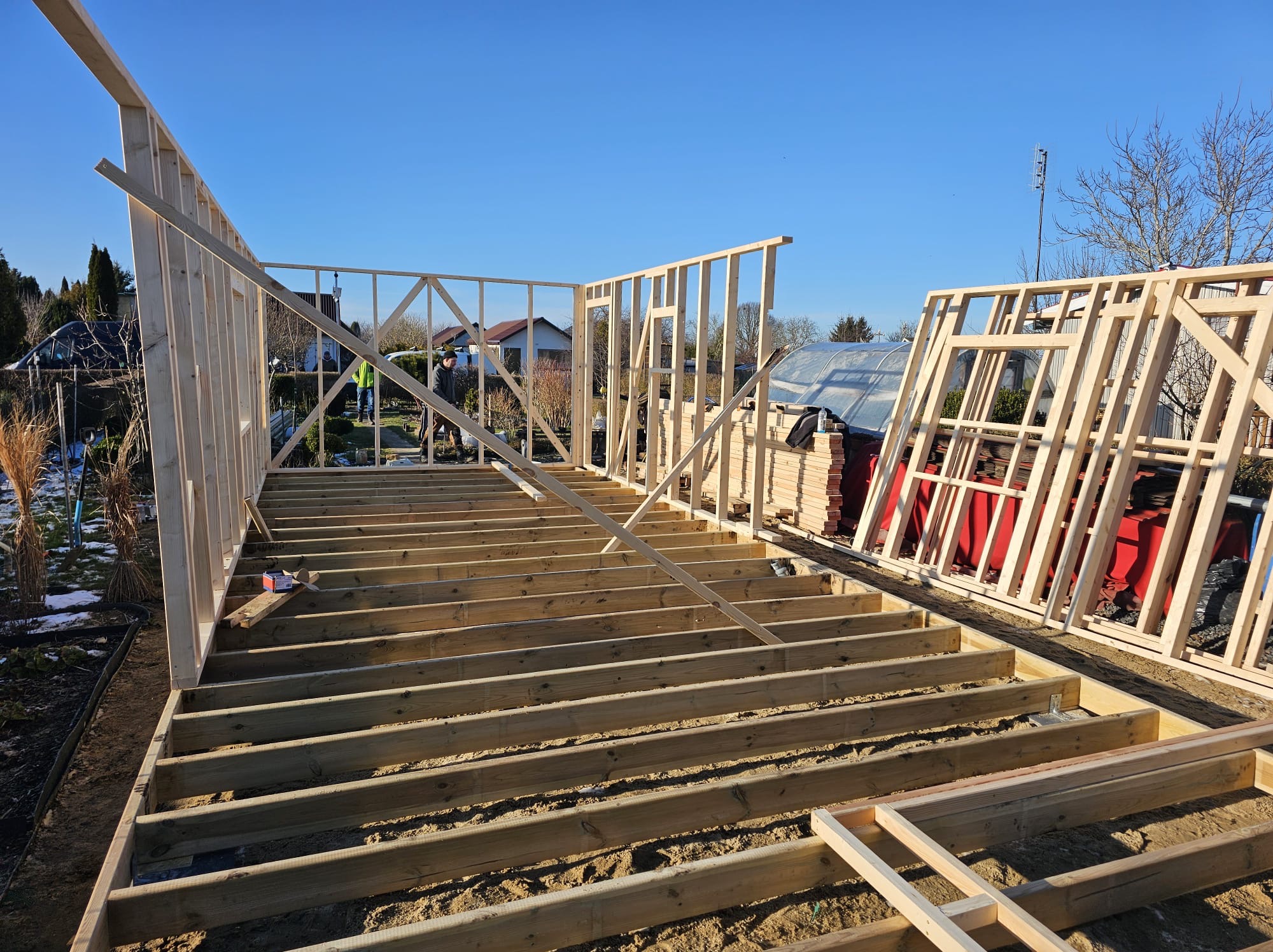 Wczesny etap budowy domu: drewniany szkielet z widoczną podłogą i fragmentami ścian, na tle ogrodu i błękitnego nieba. Widoczne detale konstrukcyjne.