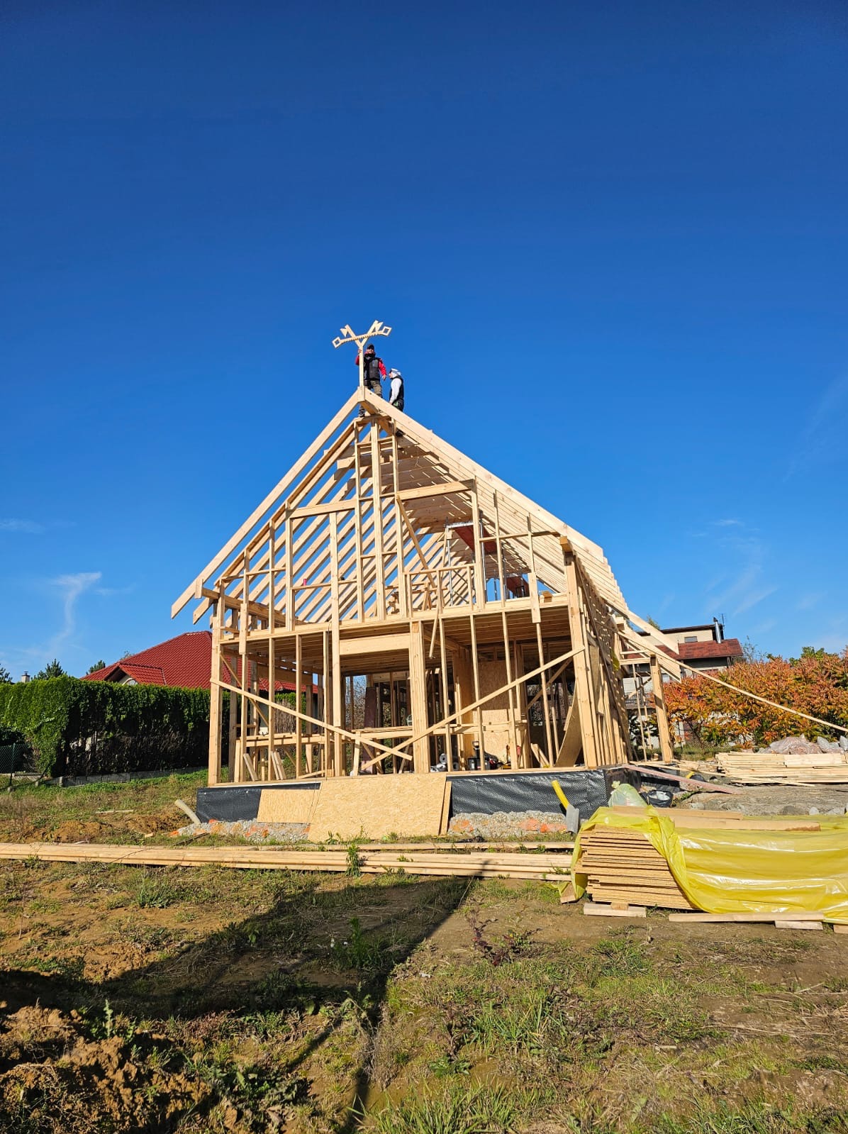 Wznoszenie konstrukcji domu z drewna, widoczna więźba dachowa i pracownicy na szczycie. Błękitne niebo w tle. Elementy konstrukcyjne na ziemi.