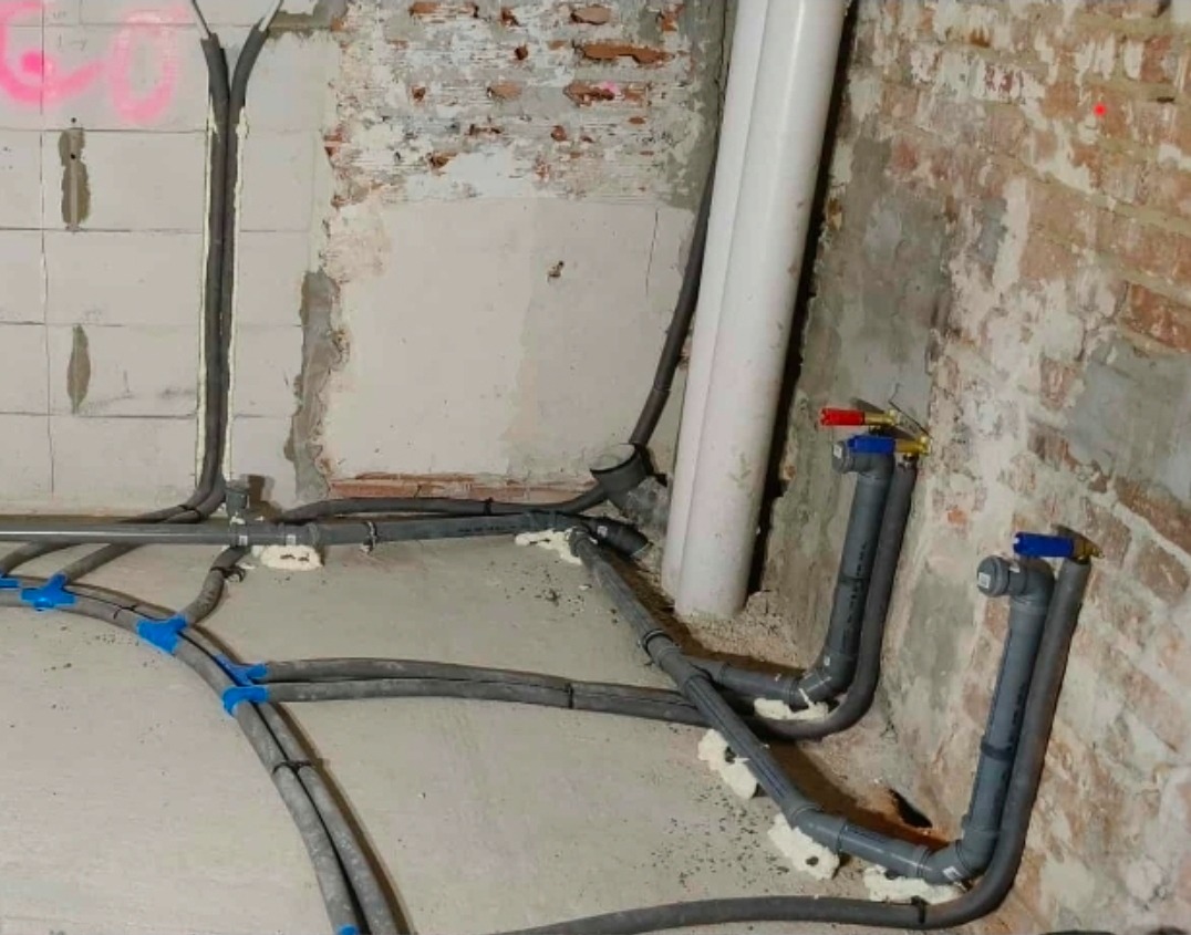 Szare rury instalacji wodnej zamocowane do ściany z cegły i bloczków, zabezpieczone pianką montażową. Widoczne niebieskie uchwyty mocujące rury do podłoża.