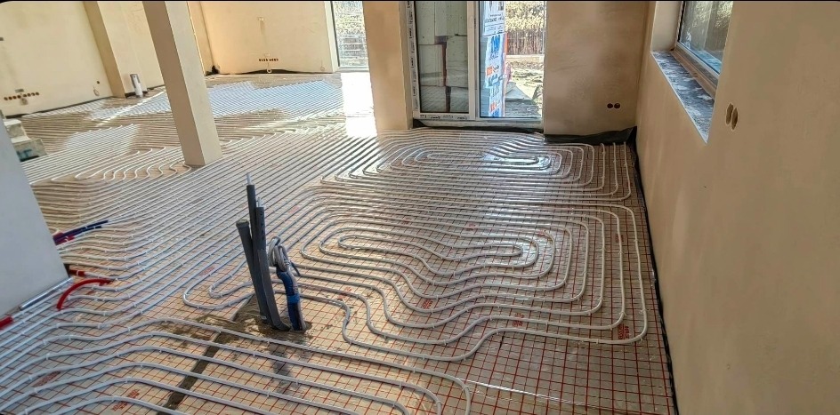 Białe rury ogrzewania podłogowego ułożone na folii z siatką w surowym wnętrzu domu. Widoczne pionowe rury instalacyjne i fragment okna.