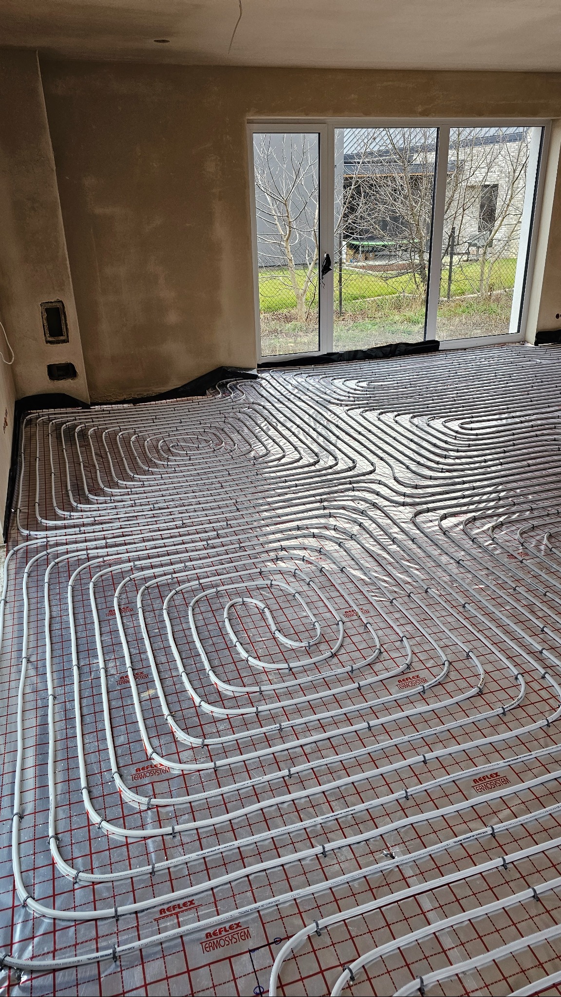 Białe rury ogrzewania podłogowego ułożone spiralnie na czerwonej macie w surowym wnętrzu z dużym oknem. Widoczne detale mocowań i napisy 'Reflex Termosystem'.