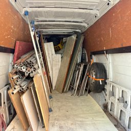 OMS Transport - Wnętrze furgonetki wypełnionej różnorodnymi przedmiotami: rurami, płytami drewnianymi, drabinką i kołem zapasowym zabezpieczonym pomarańczowym pasem transportowym.