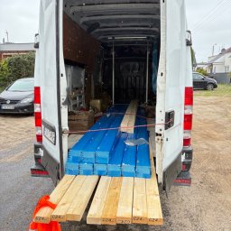 OMS Transport - Otwarty bagażnik białego busa wypełniony niebieskimi i jasnymi deskami. Na zewnątrz widoczny fragment pomarańczowej kamizelki odblaskowej i zaparkowane auta.