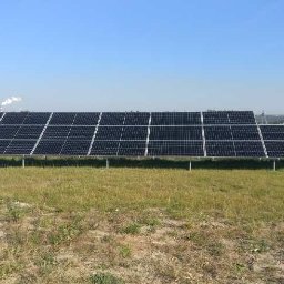 ICOM ENERGIA SPÓŁKA Z OGRANICZONĄ ODPOWIEDZIALNOŚCIĄ - Rząd paneli słonecznych na metalowych wspornikach, na tle błękitnego nieba i odległych kominów. Instalacja fotowoltaiczna na łące, widok z poziomu gruntu.