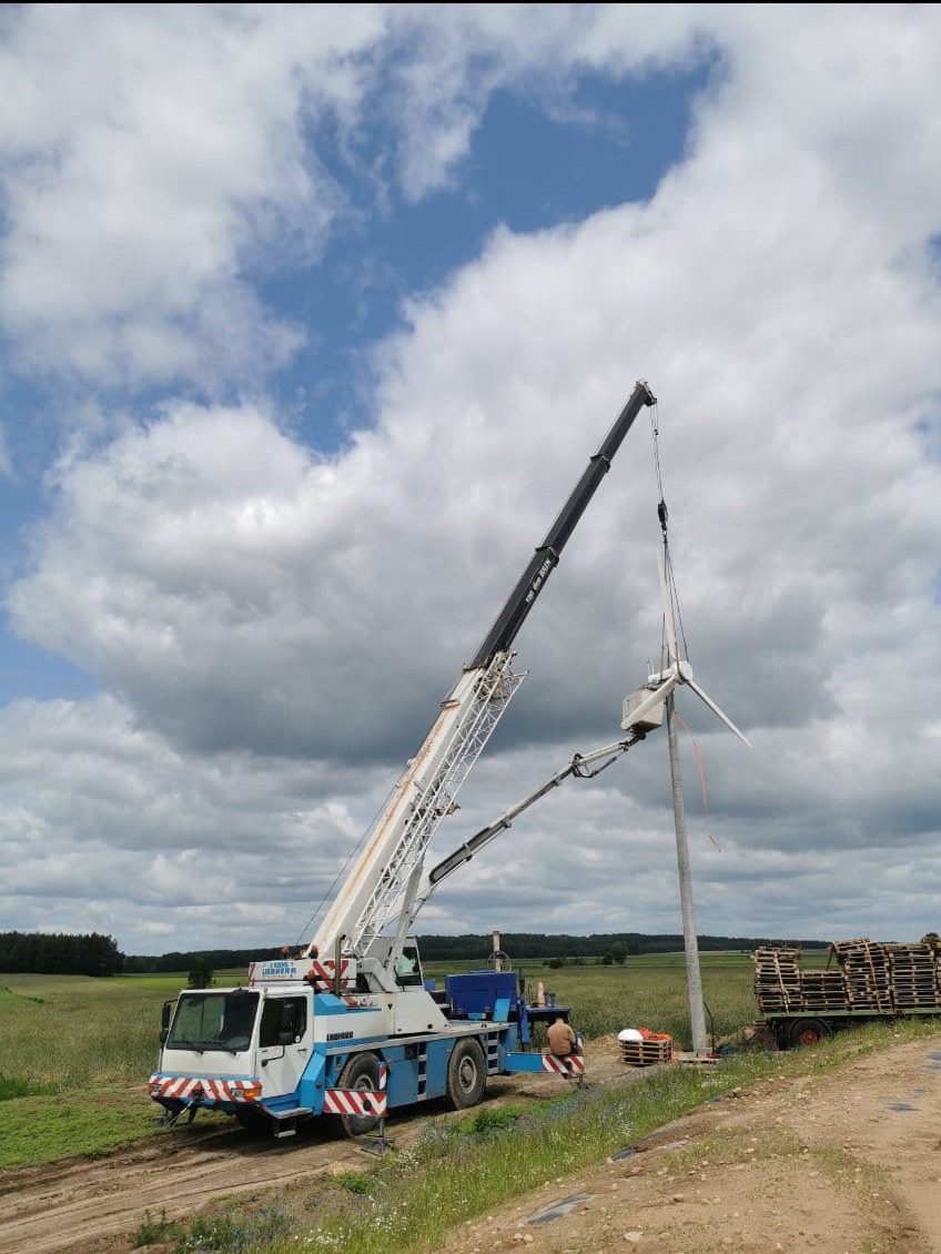 Montaż turbiny wiatrowej za pomocą dźwigu Liebherr na tle pochmurnego nieba. Widoczny pracownik w kasku i palety z materiałami. Krajobraz polny.