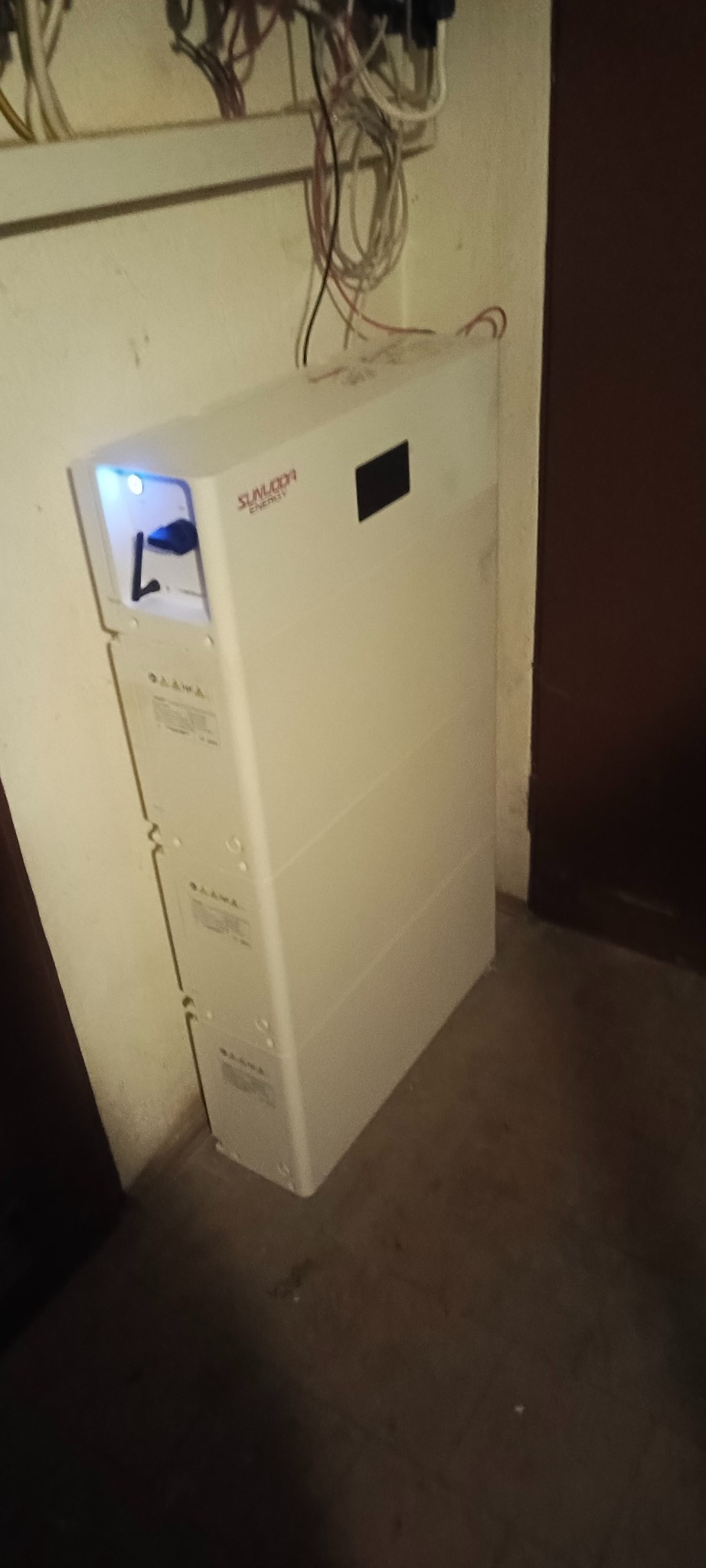 Biały moduł magazynu energii Sunwooda, ustawiony w rogu pomieszczenia, z widocznymi kablami na ścianie. Widoczne logo firmy na obudowie.